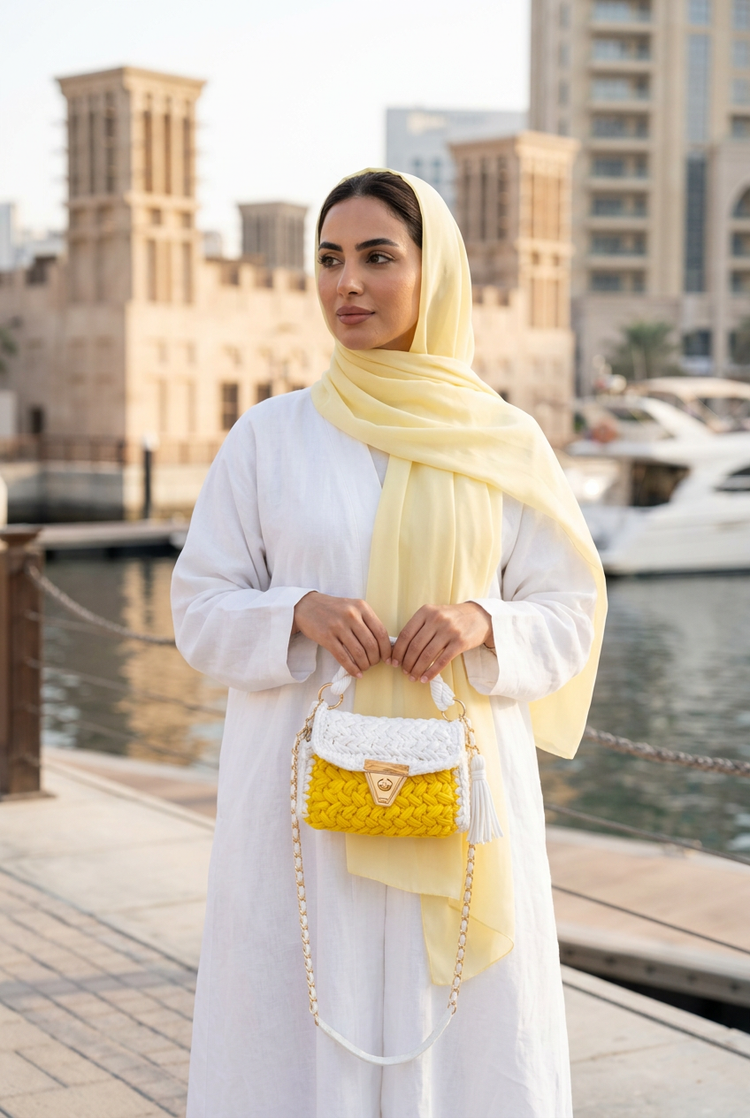 Yellow crochet top-handle bag carried with a white abaya and pale yellow shayla by the Dubai waterfront