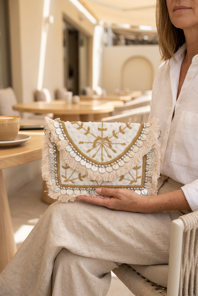 Woman in a white linen shirt holding the ivory and gold coin fringe clutch on her lap at a cafe
