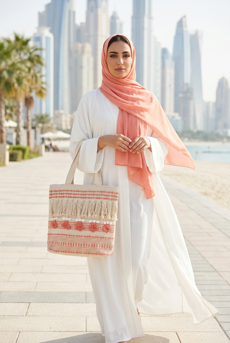 Coral floral fringe woven tote carried with a white abaya and coral shayla on a beachfront promenade