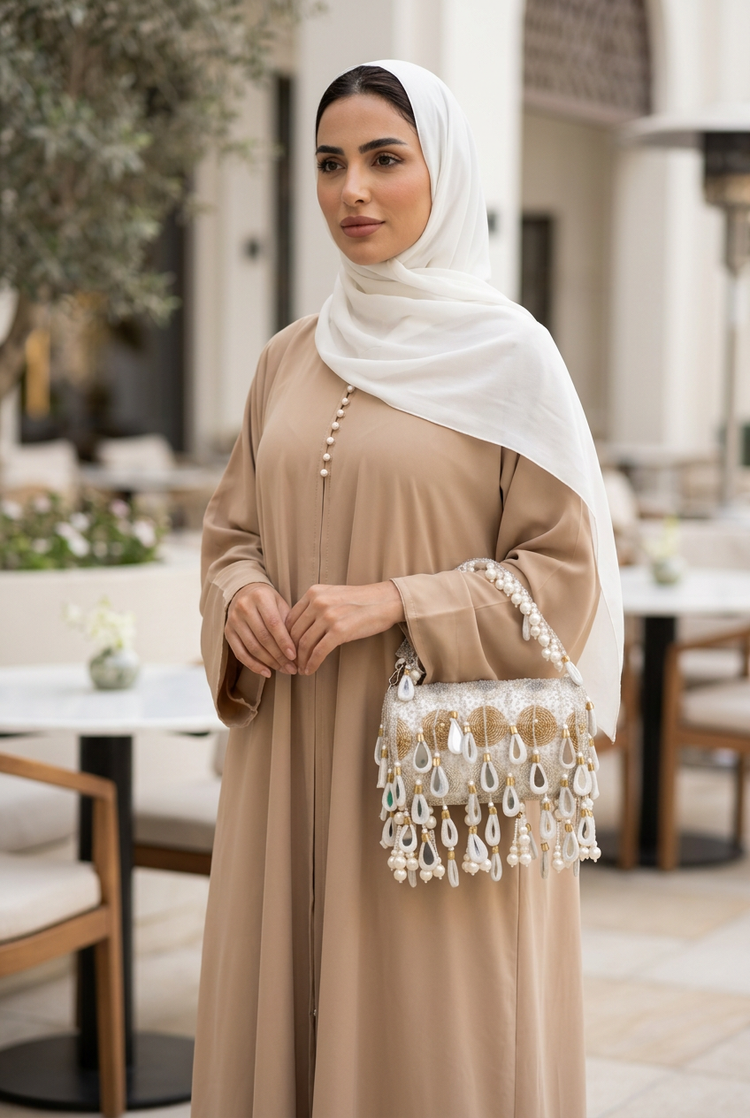 Ivory and gold teardrop pearl bag carried with a sand abaya and white shayla on a cafe terrace